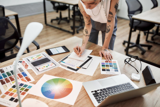 Designer workplace. Cropped view of tattooed female designer making some sketches while standing near office desk. Working with color swatch samples