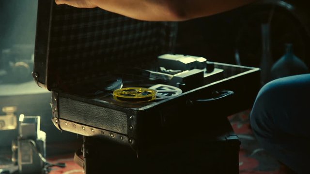 Male hands open old suitcase with vintage movie projector and film reels inside, kept in the old attic, filled with old stuff around and soft sunlight, magic atmosphere, Slow motion.