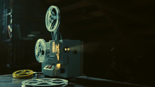 Vintage movie projector is working, showing old movie, film reels are rotating, lamp is burning inside, several film reels are lying besides, creating magic atmosphere of old cinema, Slow motion.