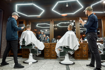 Two barbers holding mirrors and showing haircuts to father and son sitting in armchair at modern barbershop. Family, father and son visiting barber shop. Back view