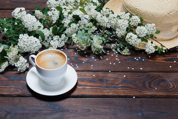 A cup of aromatic hot coffee, hat, beautiful twig of blooming White Spiraea (lat. Spiraea) isolated on dark vintage background. Concept of Spring. Space for Text. Selective focus.