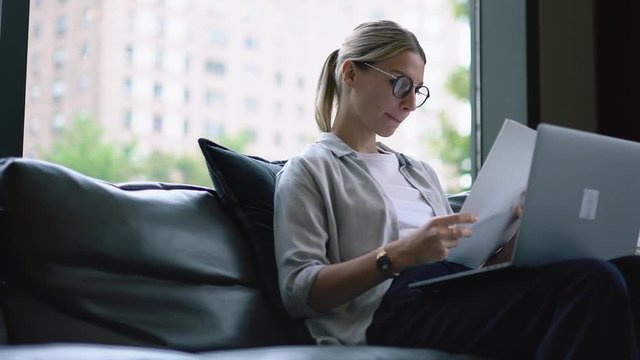 Young woman in eyeglasses reviewing paper documents and checking information during remote work at laptop in modern open space office.Hipster student doing homework and preparing coursework
