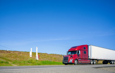 Burgundy bonnet big rig semi truck transporting cargo in dry van semi trailer driving on the road...