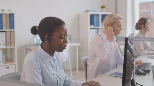 Medium shot of three diverse women wearing white shirts and headsets sitting at computer monitors in the office and consulting costumers by telephone