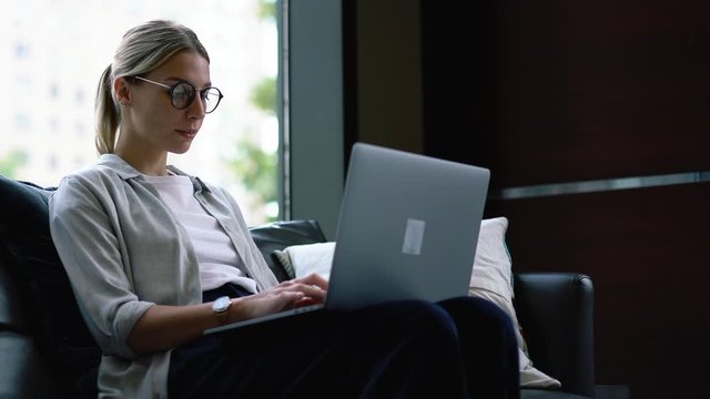 Young woman keyboarding text on laptop computer and reading information on paper documents sitting on comfortable sofa.Hipster student working on editing course work at modern netbook
