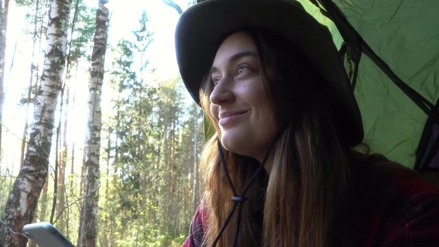 Trendy hipster traveler in hat sits in forest camping tent scrolls through her news feed smartphone checks email or texts about her adventure, looking forward and dreaming, enjoying view and solitude