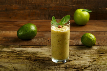 A glass of green smoothie with mint on a wooden table on a napkin near fruits and nuts.