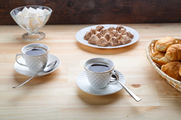 Two cups of coffee on a wooden table with croissants and sugar.