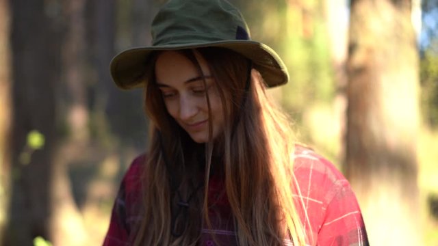 close up portrait young attractive hipster female in hat smiles and use mobile cell phone in forest camping, traveler use roaming in nature, girl looks at screen, checks social media news feed