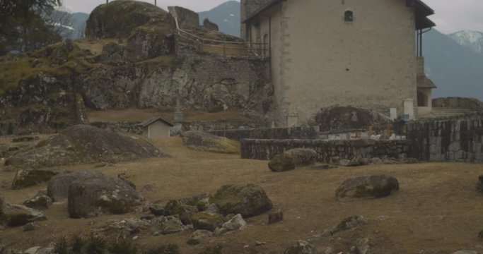 Camera tilts up towards a small rock church with three crosses alongside, in a wood of the Italian Alps 4k slow motion