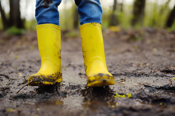 Mischievous preschooler child wearing yellow rain boots jumping in large wet mud puddle. Kid...