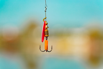 A bright spinner with a triple hook for fishing hangs on a fishing line above the water. Selective...