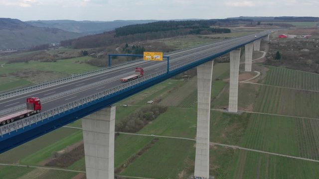 Hochmoselbr&uuml;cke High Moselle Bridge in Rhineland-Palatinate Germany with two trucks on a freeway highway cinematic inflow medium aerial drone shot on a sunny cloudy day 30p