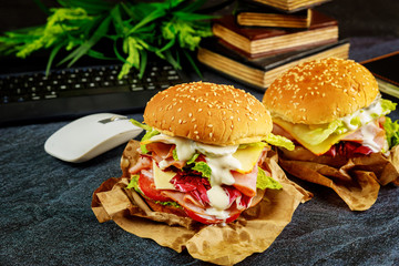 Two big sandwiches on dark desk with keyboard, mouse and books.