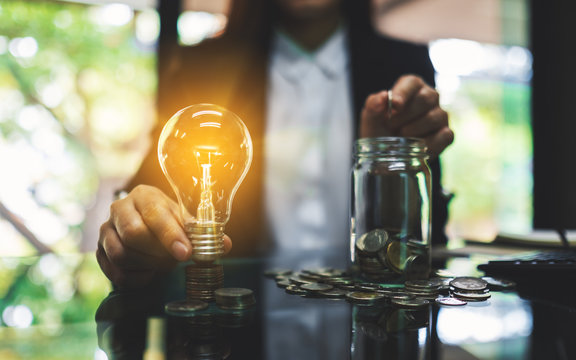 Businesswoman holding a light bulb over coins stack on the table while putting coin into a glass jar for saving energy and money concept
