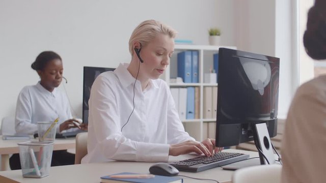 Pan shot of Caucasian blond-haired female customer service worker searching information for client in electronic database while her female coworkers working on background