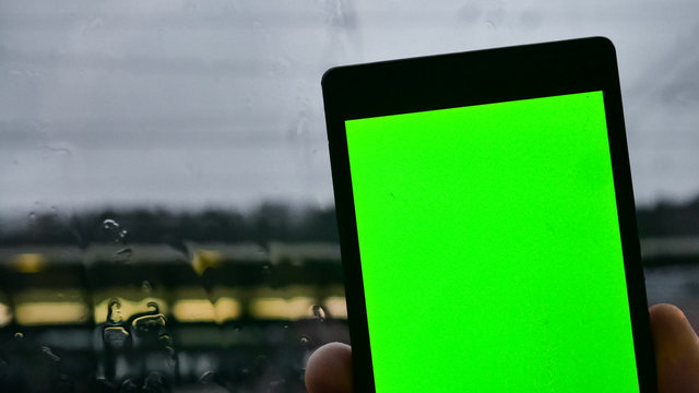 Green screen, phone. A man holds a phone in his hand with a green screen against the background of a window wet from the rain. 