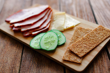 Slicing ham, cucumbers and cheese with ruddy thin breads lie on a wooden board.