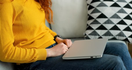 Naklejka premium Close up of redhead girl using silver laptop while sitting on sofa in living room at home.