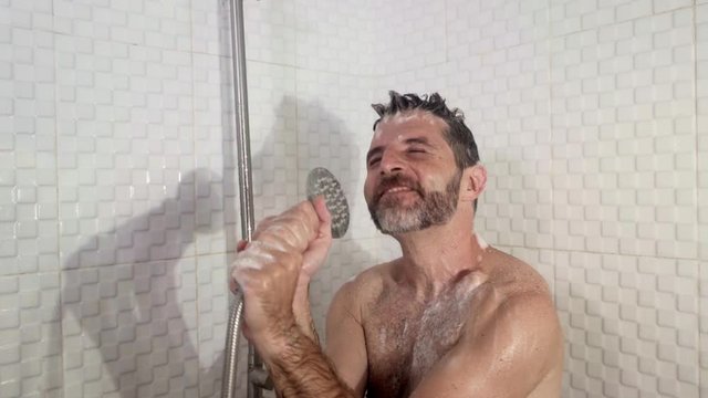  funny shot of young attractive and happy man with beard dancing and singing in the shower carefree and crazy at home pretending holding a microphone enjoying