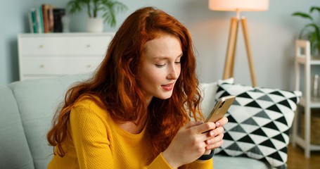 Portrait of beautiful cheerful redhead girl relax sit on sofa at home using cellphone.