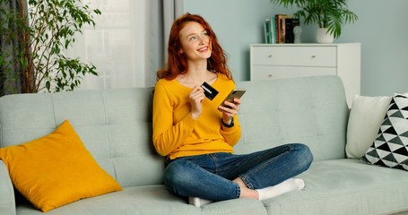 Young beautiful red-haired woman sitting on couch in living room and shopping online on mobile phone.