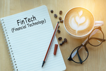 Concept fintech message on notebook with glasses, pencil and coffee cup on wooden table.