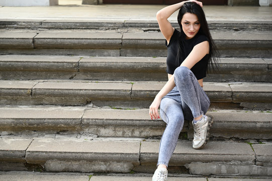 Full-length photo in spring weather outdoors, caucasian brunette girl with a slim figure in jeans and a black T-shirt. Fashionable model sits on the stairs of the building.
