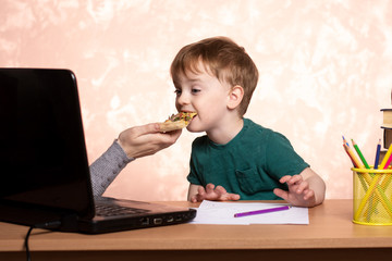 a three-year-old child sits at a table with a laptop and books and eats a slice of pizza from her mother&rsquo;s hands. quarantined classes online. difficulties of distance learning.