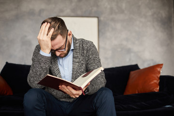 Young businessman holding his head while sitting on sofa and reading his plans and tasks in note...