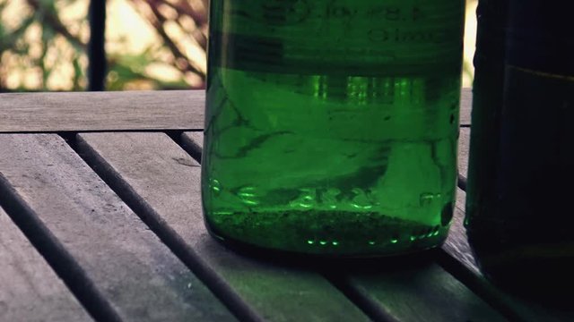 The bottom of a selection of green glass bottles on a wooden table, sideways movement with railings and a blurred lush garden background
