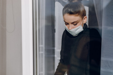 young attractive girl in a protective medical mask looks out the window. isolation during the epidemic. home isolation.