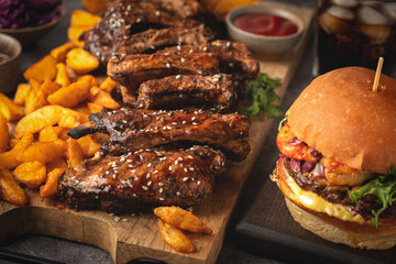 Barbecue pork ribs on a wooden board, potato wedges, burger and cola glass, sause. Fast food. Close-up.