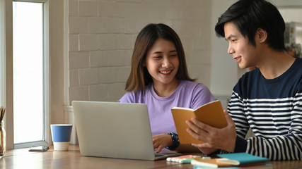 Couple tutoring their lesson while sitting together in front of computer laptop at the wooden working desk that surrounded by coffee cup, stack of notebook, marker pens and smartphone.