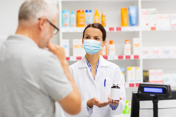 Fototapeta na wymiar medicine, pharmaceutics, health care and people concept - apothecary wearing face protective medical mask for protection from virus disease showing drug to senior man customer at drugstore