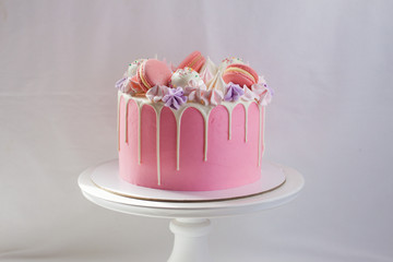 Tender pink cake decorated with melted white chocolate, macaroons, meringues, cake pops and candies. Plain background.