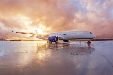 Passenger commercial wide-body aircraft parked with a wet apron at the airport during evening rain at sunset with beautiful sky.