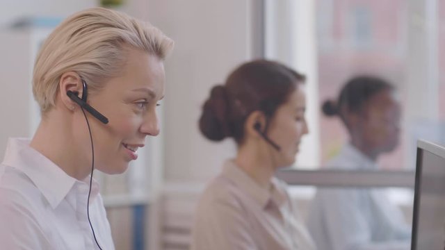 Close up of Caucasian blond woman wearing white shirt and headset giving phone advice to interlocutor and then looking at camera while two diverse operators working on background