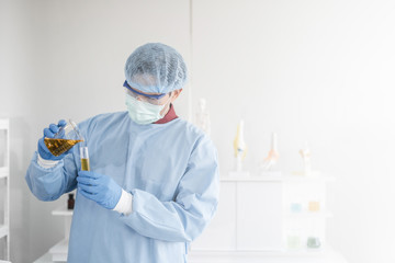 A male scientist with black hair wearing a blue protective lab coat, glove,  mask, and protective glassware pouring liquid solution from a beaker into a test tube in a laboratory setting.