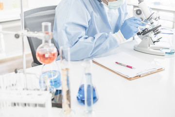 A scientist wearing a blue protective lab coat, glove, and mask looking into a microscope in a laboratory setting with test tubes.