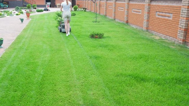 A woman mows grass with a lawn mower in the backyard of a house. To work in the garden. Back view.