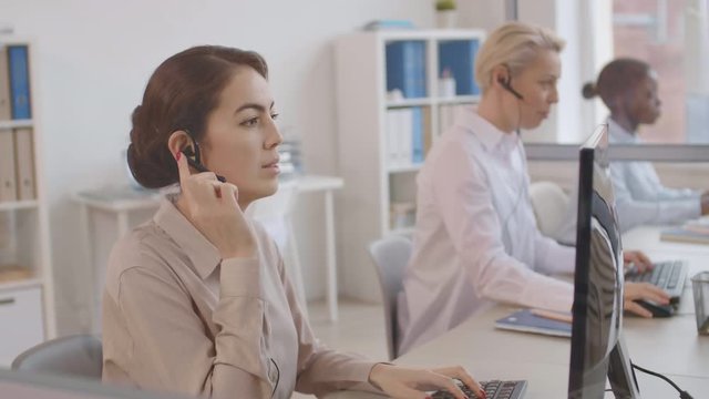 Medium shot of three multiethnic female operators wearing white shirts and headphones sitting at computer displays in the office and consulting interlocutors by phone