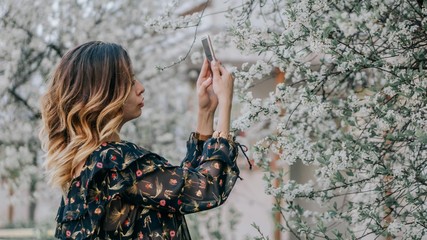 smiling girl photographs a blossoming cherry on a smartphone. young girl takes a selfie near sakura