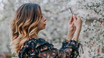 smiling girl photographs a blossoming cherry on a smartphone. young girl takes a selfie near sakura