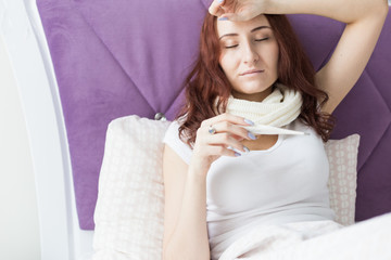 An attractive young brunette woman sits in her bed with a thermometer. Colds and home treatment