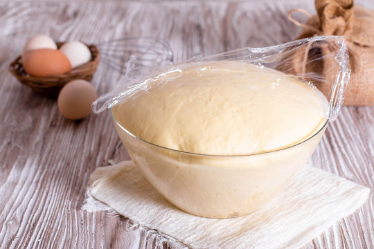 Yeast dough in a glass bowl under cling film on wooden table