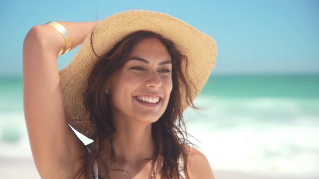Close up face of young stylish woman wearing straw hat enjoying the breeze at beach. Happy tanned latin woman laughing during summer holiday. Beautiful fashionable girl relaxing at beach having fun.