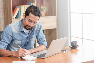 Fototapeta na wymiar 30s handsome businessman take a short note on paper while reading article from internet. Modern arab businessman in casual clothes working at home.