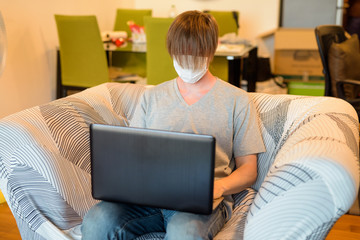 Young man with long hair wearing mask and looking shabby while using laptop in quarantine at home