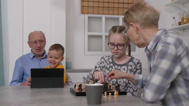 Close-up of focused granddaughter learning to play chess with grandma while little grandson together with granddad enjoying cartoons on tablet. Family of different ages spending free time in kitchen